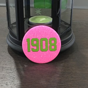 Op de afbeelding: Een ronde, roze glitterknop met het nummer "1908" in groen. De knop bevindt zich voor een zwarte lantaarn met een brandende groene kaars erin. De knop ligt op een donker houten oppervlak.