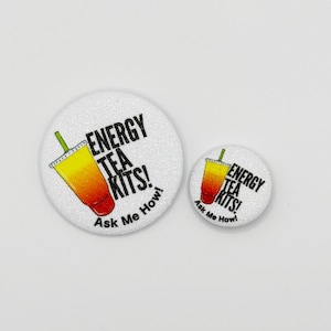 May include: Two white glittery round buttons with the text "ENERGY TEA KITS! Ask Me How!" and a graphic of a yellow and red drink. One button is larger than the other.
