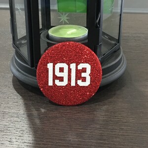 May include: A round, red glitter button with the year "1913" in white block letters. The button is resting on a dark wooden surface near a black lantern with a lit green candle inside.