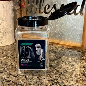 Op de afbeelding: Een doorzichtige plastic pot met een zwarte deksel, met een etiket met de tekst "CR7 DRIVE" en een portret van een man. De pot is open, waardoor de inhoud zichtbaar is. De achtergrond bevat een decoratief metalen bord.