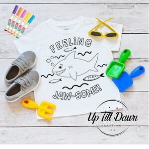 Pode incluir: Camiseta branca com um contorno preto e branco de um tubar&atilde;o e o texto "Feeling Jaw-some!"  A camiseta est&aacute; pronta para ser colorida com marcadores. A imagem tamb&eacute;m inclui um par de &oacute;culos de sol amarelos, duas p&aacute;s de pl&aacute;stico e um ancinho de pl&aacute;stico amarelo.