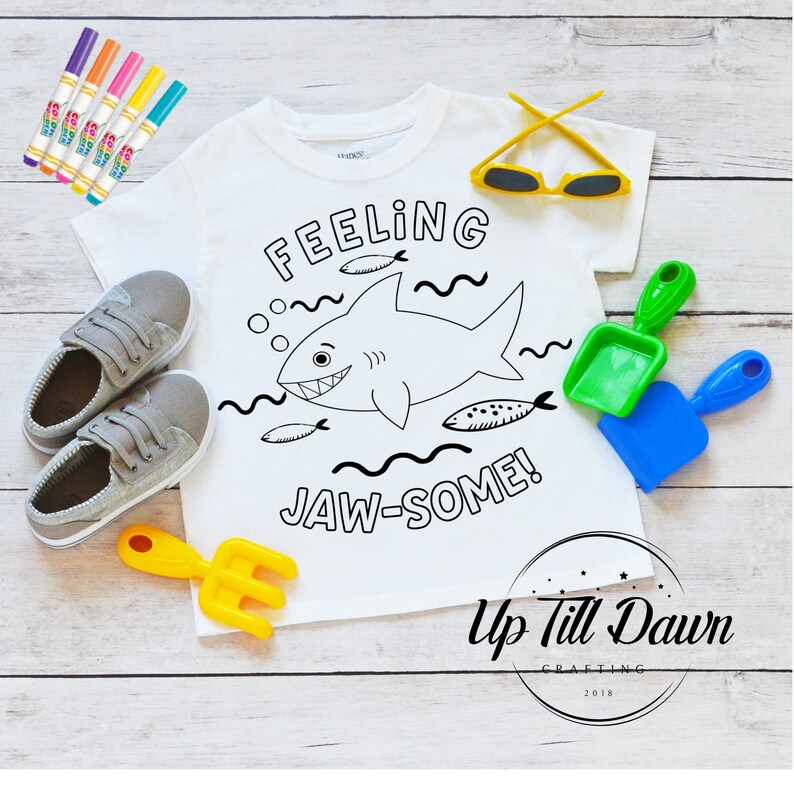 Pode incluir: Camiseta branca com um contorno preto e branco de um tubar&atilde;o e o texto "Feeling Jaw-some!"  A camiseta est&aacute; pronta para ser colorida com marcadores. A imagem tamb&eacute;m inclui um par de &oacute;culos de sol amarelos, duas p&aacute;s de pl&aacute;stico e um ancinho de pl&aacute;stico amarelo.