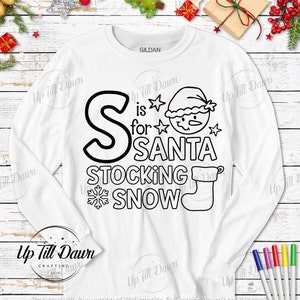 Op de afbeelding: Een wit longsleeve shirt met een zwart-wit ontwerp dat "S is for SANTA STOCKING SNOW" zegt. Het ontwerp bevat een sneeuwpop, een sok en een sneeuwvlok.