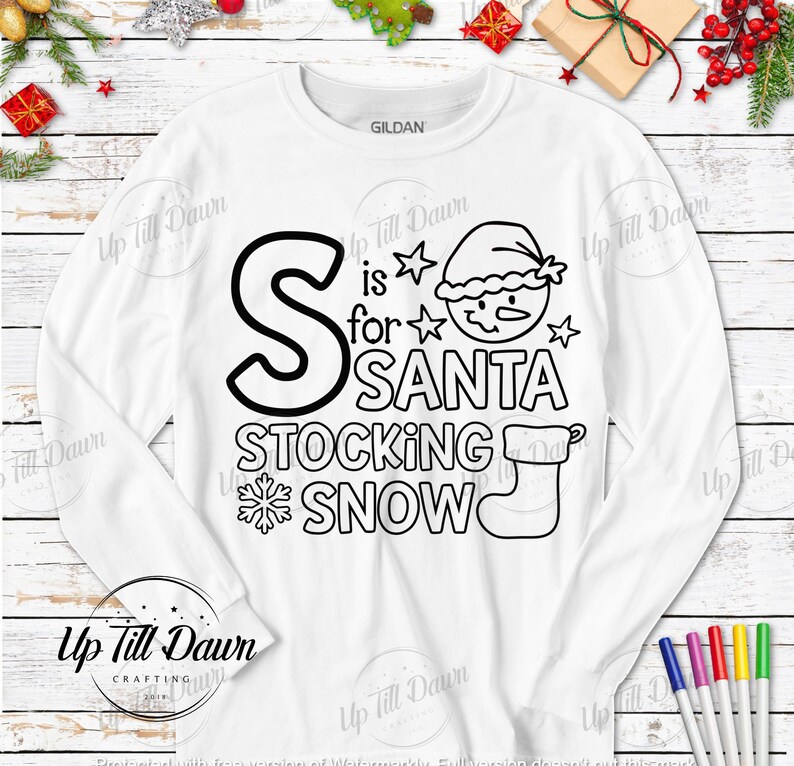 Op de afbeelding: Een wit longsleeve shirt met een zwart-wit ontwerp dat "S is for SANTA STOCKING SNOW" zegt. Het ontwerp bevat een sneeuwpop, een sok en een sneeuwvlok.