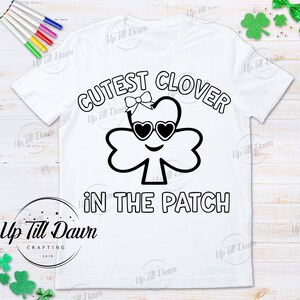 Puede incluir: Una camiseta blanca con un dise&ntilde;o en blanco y negro de un tr&eacute;bol con gafas de sol y un lazo. El texto "CUTEST CLOVER IN THE PATCH" est&aacute; escrito alrededor del tr&eacute;bol.