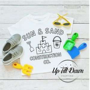 May include: White t-shirt with a black and white design that says "Sun & Sand Construction Co." with a sandcastle, bucket, and shovel. The design is in a circle with a yellow star at the top. The shirt is on a wooden surface with a yellow rake, blue shovel, and green shovel.