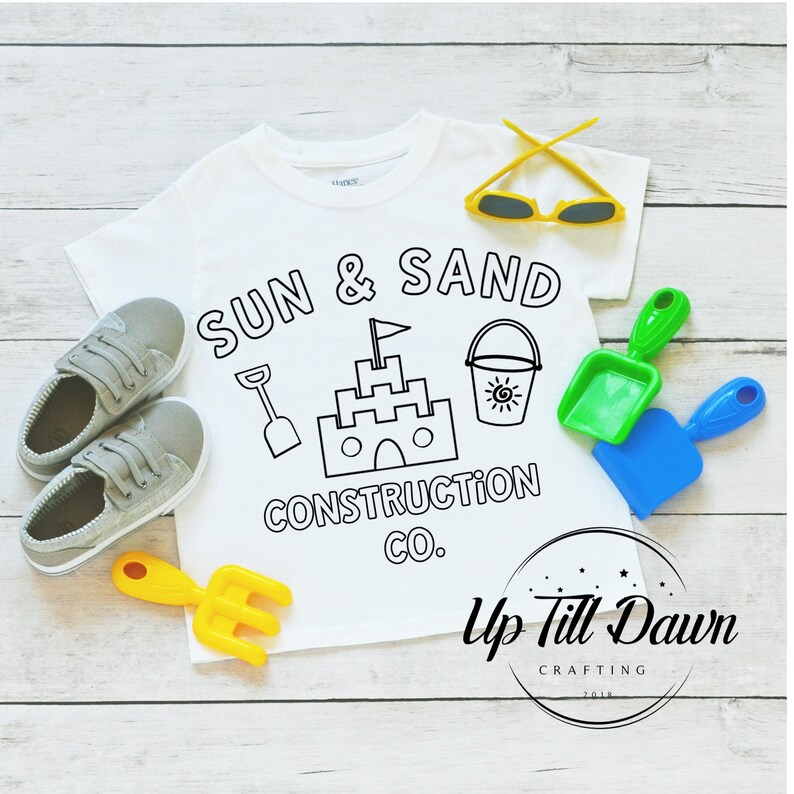 May include: White t-shirt with a black and white design that says "Sun & Sand Construction Co." with a sandcastle, bucket, and shovel. The design is in a circle with a yellow star at the top. The shirt is on a wooden surface with a yellow rake, blue shovel, and green shovel.