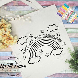May include: A cream-colored tote bag with the phrase "Follow The Rainbow" and a rainbow graphic. The design includes stars and clouds. Several colorful markers are visible on the right side of the bag.