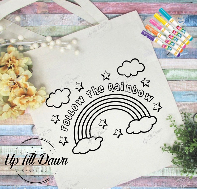 May include: A cream-colored tote bag with the phrase "Follow The Rainbow" and a rainbow graphic. The design includes stars and clouds. Several colorful markers are visible on the right side of the bag.