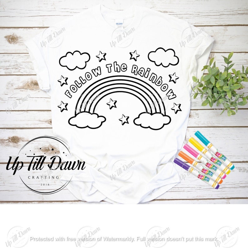 May include: White t-shirt with a black outline design featuring a rainbow, clouds, and stars, with the text "Follow The Rainbow." The shirt is accompanied by a set of colorful markers, perfect for coloring the design.