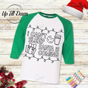 Op de afbeelding: Een wit en groen kleur-je-eigen kerstthema t-shirt met de tekst "Ik kan niet slapen, Sinterklaas komt" en een afbeelding van een glas melk, een koekje en een ingepakt cadeau.