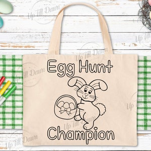 May include: A white canvas tote bag with black outlines of a bunny holding a basket of eggs. The text "Egg Hunt Champion" is printed on the bag.