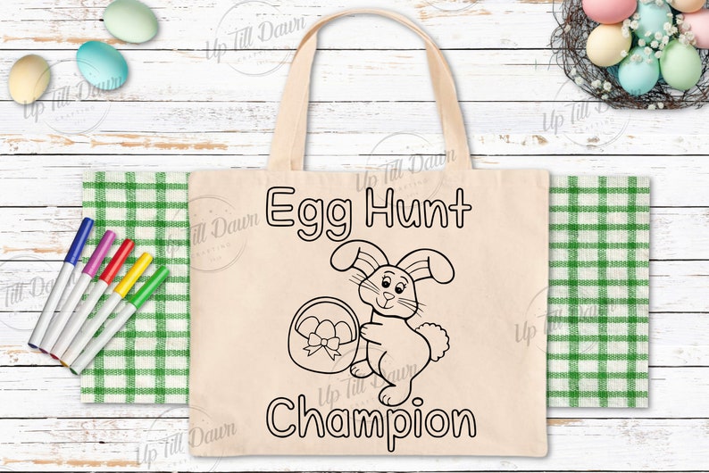 May include: A white canvas tote bag with black outlines of a bunny holding a basket of eggs. The text "Egg Hunt Champion" is printed on the bag.