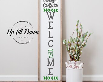 Könnte beinhalten: Ein Holzschild mit den Worten "Ich hoffe, du hast Kaffee mitgebracht" und "Willkommen" mit einer Kaffeetassengrafik dazwischen. Das Schild ist mit grünen Blättern verziert.