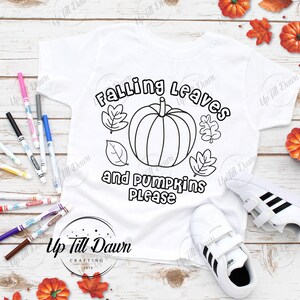 Puede incluir: Una camiseta blanca con un contorno en blanco y negro de una calabaza y hojas. El texto "Falling Leaves and Pumpkins Please" est&aacute; escrito en negro. La camiseta est&aacute; sobre una superficie de madera con hojas de oto&ntilde;o.