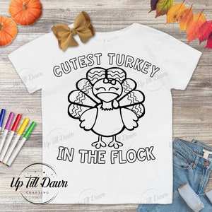 May include: A white t-shirt with a black and white outline of a turkey with a bow on its head. The text "CUTEST TURKEY IN THE FLOCK" is printed on the shirt.