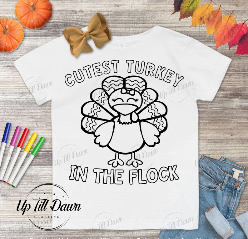 May include: A white t-shirt with a black and white outline of a turkey with a bow on its head. The text "CUTEST TURKEY IN THE FLOCK" is printed on the shirt.