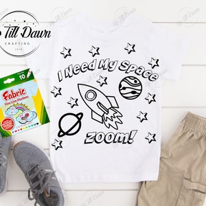May include: A white t-shirt with a black and white design of a rocket ship, stars, and planets. The text "I Need My Space" and "zoom!" are also included in the design. The shirt is lying on a white surface with a pack of fabric markers.