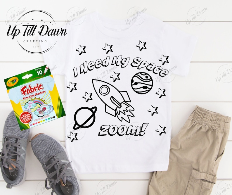 May include: A white t-shirt with a black and white design of a rocket ship, stars, and planets. The text "I Need My Space" and "zoom!" are also included in the design. The shirt is lying on a white surface with a pack of fabric markers.