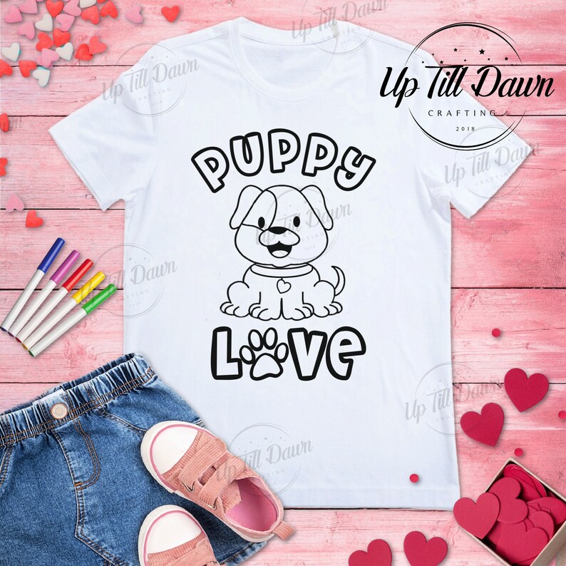 May include: A white t-shirt with a black and white outline of a puppy and the words "PUPPY LOVE" in black. The shirt is on a pink wooden surface with a pair of jeans, pink shoes, and red hearts.