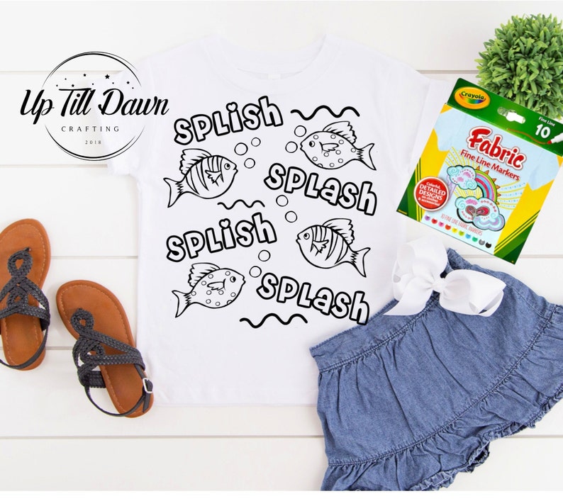 Pode incluir: Uma camiseta branca com um contorno preto de peixes e as palavras "Splish Splash" repetidas. A camiseta est&aacute; sobre uma superf&iacute;cie branca com um par de sand&aacute;lias marrons e um pacote de marcadores Crayola Fabric Fine Line.
