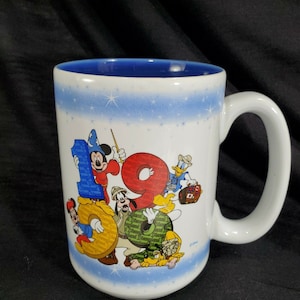 May include: White ceramic mug with a blue interior and handle. The mug features the number "19" with Disney characters Mickey Mouse, Minnie Mouse, Donald Duck, Goofy, and Pluto. The mug has a light blue band with white stars.
