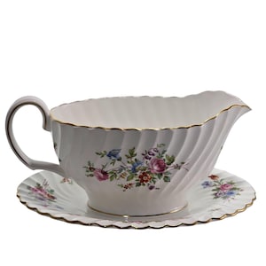 May include: A white porcelain gravy boat and saucer set. The boat features a handle and a scalloped rim, adorned with a floral pattern in pink, blue, and green. Gold trim edges the pieces. The saucer echoes the boat's design.