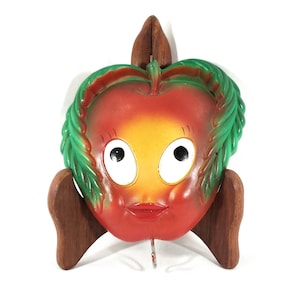 May include: A red and yellow ceramic apple with a green leaf design and a smiling face. The apple is on a wooden stand.