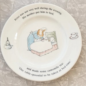 May include: A white ceramic plate with a picture of Peter Rabbit in bed. The plate has text that reads "Peter was not very well during the evening. His mother put him to bed, and made some camomile tea: 'One table-spoonful to be taken at bed-time.'"