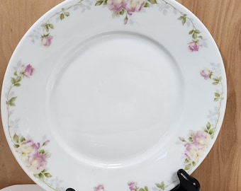 May include: A round white ceramic dinner plate featuring a delicate floral border pattern. The rim is decorated with small pink rose blossoms, green leaves, and thin trailing vines that encircle the edge of the plate against a plain white center.