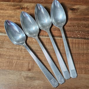 May include: Four silver spoons with oval bowls and long, ridged handles. The spoons are arranged diagonally on a wooden surface, showcasing their polished, reflective surfaces. The handles have a geometric design at the base.