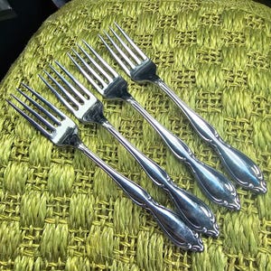 May include: Four silver forks with ornate handles are arranged diagonally on a textured, light green surface. The forks have tines and a decorative pattern on the handles. The silverware is likely for a formal dining setting.