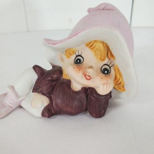 May include: A ceramic figurine of a whimsical elf with a pink hat and a burgundy outfit. The elf has blonde hair, rosy cheeks, and is resting its chin on its hand. The figurine is lying down.