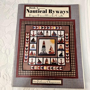 May include: A book titled "Nautical Byways" featuring a quilt pattern with nautical themes. The cover displays a quilt design with sailboats, lighthouses, and anchors in a patchwork style, framed by a red and black checkered border.
