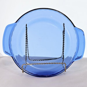 May include: A vintage, cobalt blue glass casserole dish with two handles. The dish is round with a slightly scalloped edge. It is displayed on a decorative metal stand. The dish is empty and appears to be in good condition.