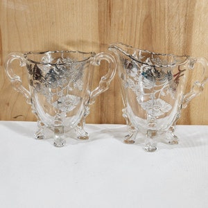 May include: A set of two clear glass sugar and creamer with silver floral designs and three legs.