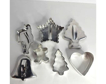 May include: A collection of seven silver-colored metal cookie cutters in various shapes, including a star, a Christmas tree, a bell, a heart, a gingerbread person, and two other distinct forms, all featuring smooth edges and metallic surfaces.