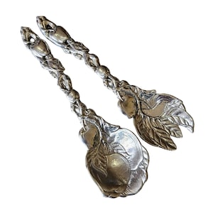 Może przedstawiać: Para srebrnych przyborów do serwowania. Uchwyty mają szczegółowy wzór liści i małych okrągłych kształtów. Końcówki do serwowania mają kształt jabłek z liśćmi. Przybory służą prawdopodobnie do serwowania sałatek lub innych potraw.