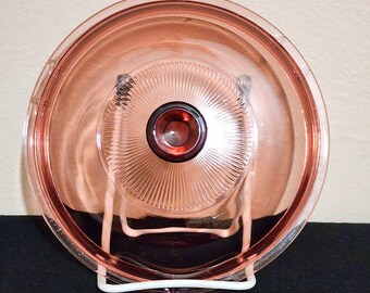 May include: A round, transparent pink glass lid featuring a central knob with a dark red center. The lid has a radiating ribbed pattern extending from the center to the outer edge, and it is supported by a white wire stand for display purposes.