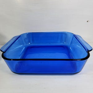 May include: A square, cobalt blue glass baking dish with a black trim. The dish has a rectangular shape with slightly flared sides and handles on each end. The dish is empty and sits on a white surface.