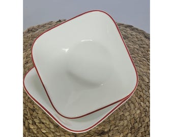 May include: A set of two square white ceramic bowls with rounded corners and a thin red rim along the edges. The bowls are stacked on top of each other, featuring a smooth, glossy finish and a deep, concave center for holding food items.