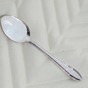 May include: A silver spoon with a rounded bowl and a decorative handle. The spoon is lying on a white fabric surface.