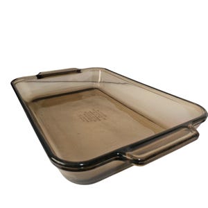 May include: An amber-tinted rectangular glass baking dish with two handles. The dish has a slightly curved shape and is made of transparent glass. The interior of the dish has a faint, etched text. Suitable for baking and serving.
