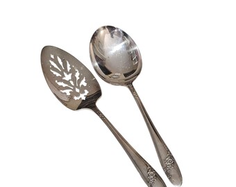 May include: A silver-toned serving set consisting of a slotted server and a solid spoon. Both utensils feature a polished finish and a decorative floral pattern embossed along the handles. The server has a leaf-shaped cutout design.