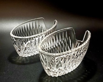 May include: Two clear, crystal glass serving dishes. Each dish has a boat-like shape with a handle on one side and a textured, ridged pattern. The dishes are on a dark surface.