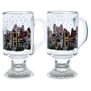May include: Two clear glass mugs with a winter village scene printed on them. The mugs have a footed base and a handle. The scene includes houses, trees, and snow.