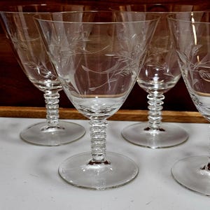 May include: A set of clear, stemmed wine glasses. Each glass features a delicate etched floral design on the bowl. The stems have a textured, beaded appearance, and the bases are round and flat. The glasses are arranged on a light surface.
