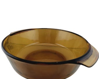 May include: A round, translucent amber-colored glass bowl featuring a smooth, polished finish and a single integrated handle extending from one side. The bowl has a wide, open top and a curved base, constructed from a single piece of tinted glass.