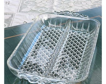 May include: A rectangular clear glass serving dish featuring a central divider, a textured hobnail pattern on the base, vertical ribbed sides, and decorative scrollwork handles on both ends. The dish is transparent and has a polished finish.