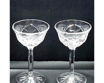 May include: Two clear glass stemmed goblets featuring a detailed cut glass pattern on the bowls and a starburst design on the circular bases. The glassware has a wide rim and a slender stem, showcasing intricate geometric etchings throughout.